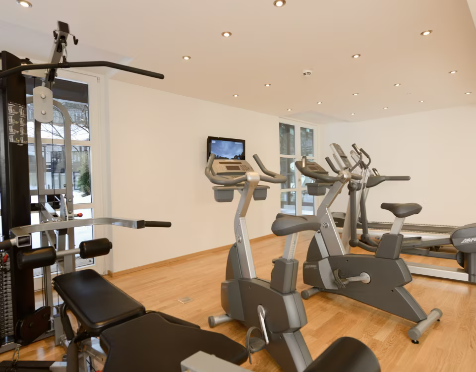 Ein heller Fitnessraum mit mehreren Heimtrainern, einem Laufband und einem Krafttrainingsgerät auf Holzboden.