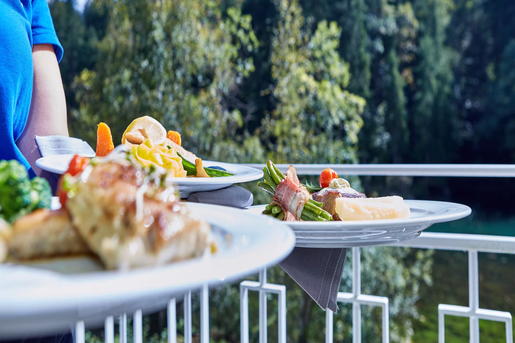 Servierperson bringt Teller mit verschiedenen Gourmetgerichten auf einem Balkon mit grünem Wald im Hintergrund.