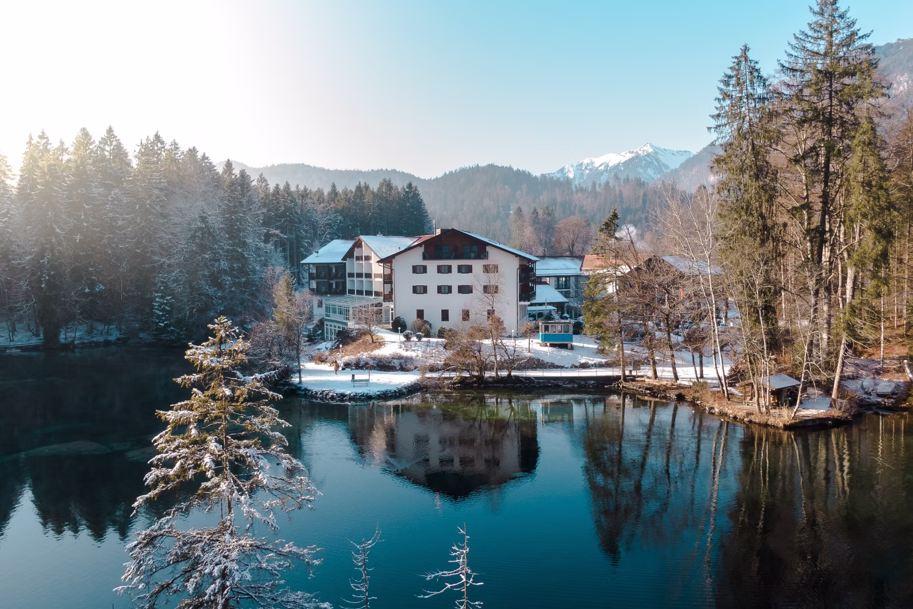 Verschneites Gebäude am Ufer eines ruhigen Sees mit Spiegelung, umgeben von Bäumen und Bergen im Hintergrund.