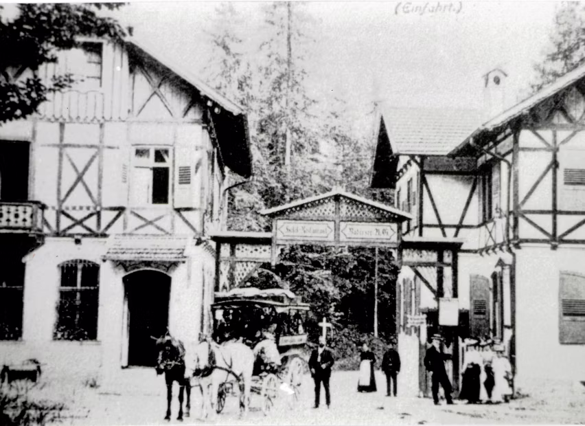 Schwarz-weiß-Foto von Fachwerkhäusern mit einem Pferdewagen und mehreren Menschen an einem Eingangstor in einer bewaldeten Gegend.