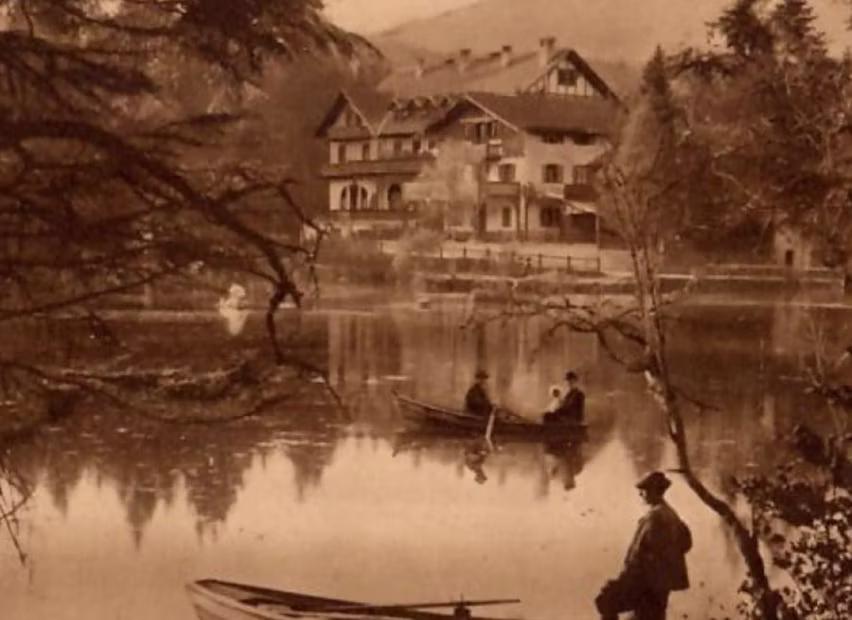 Sepiafarbenes Bild eines ruhigen Sees mit zwei Booten, einem Mann im Vordergrund am Ufer und Häusern im Hintergrund.