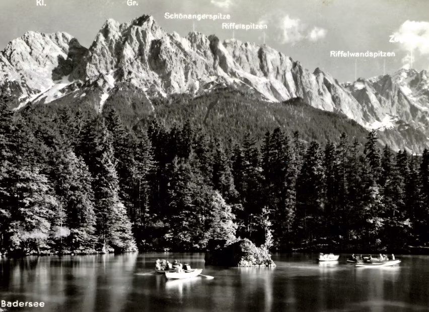 Schwarzweiß-Foto von Badersee mit mehreren Ruderbooten auf ruhigem Wasser, umgeben von Nadelwald und im Hintergrund markante Berggipfel mit Beschriftungen.