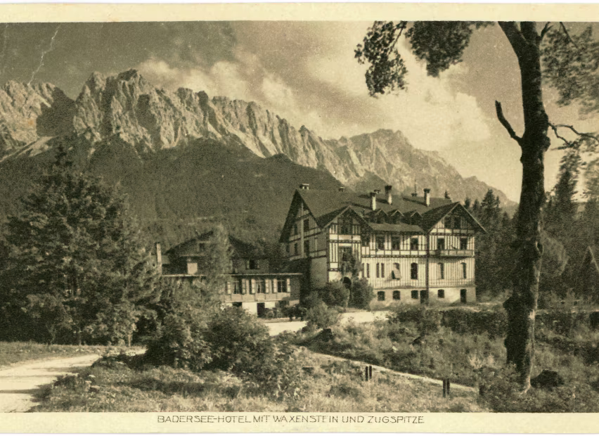 Schwarz-weiß Foto eines großen Hotels im Alpenstil mit Bergen im Hintergrund und Bäumen im Vordergrund.