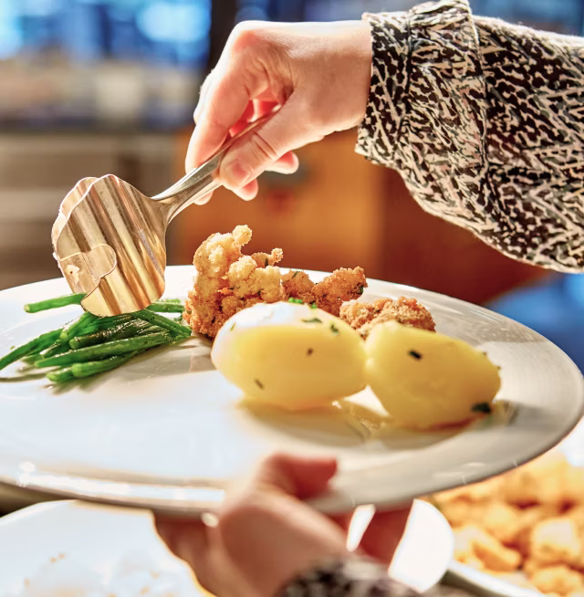 Person legt mit Zange grüne Bohnen auf einen Teller mit zwei Kartoffeln und paniertem Fleisch.