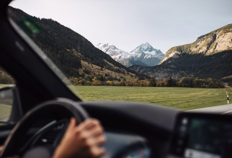 Blick aus einem Auto auf eine grüne Wiese, umgeben von bewaldeten Hügeln und schneebedeckten Bergen im Hintergrund.