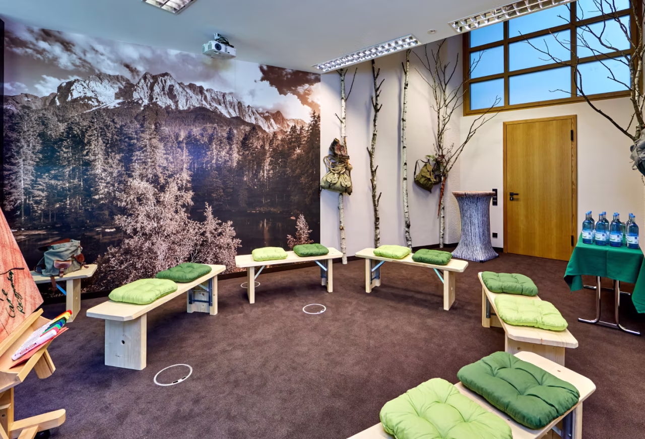 Raum mit Sitzbänken in Halbkreis, grünen Kissen, großen Fototapeten von Bergen und Bäumen, und Dekoration mit Birkenästen.
