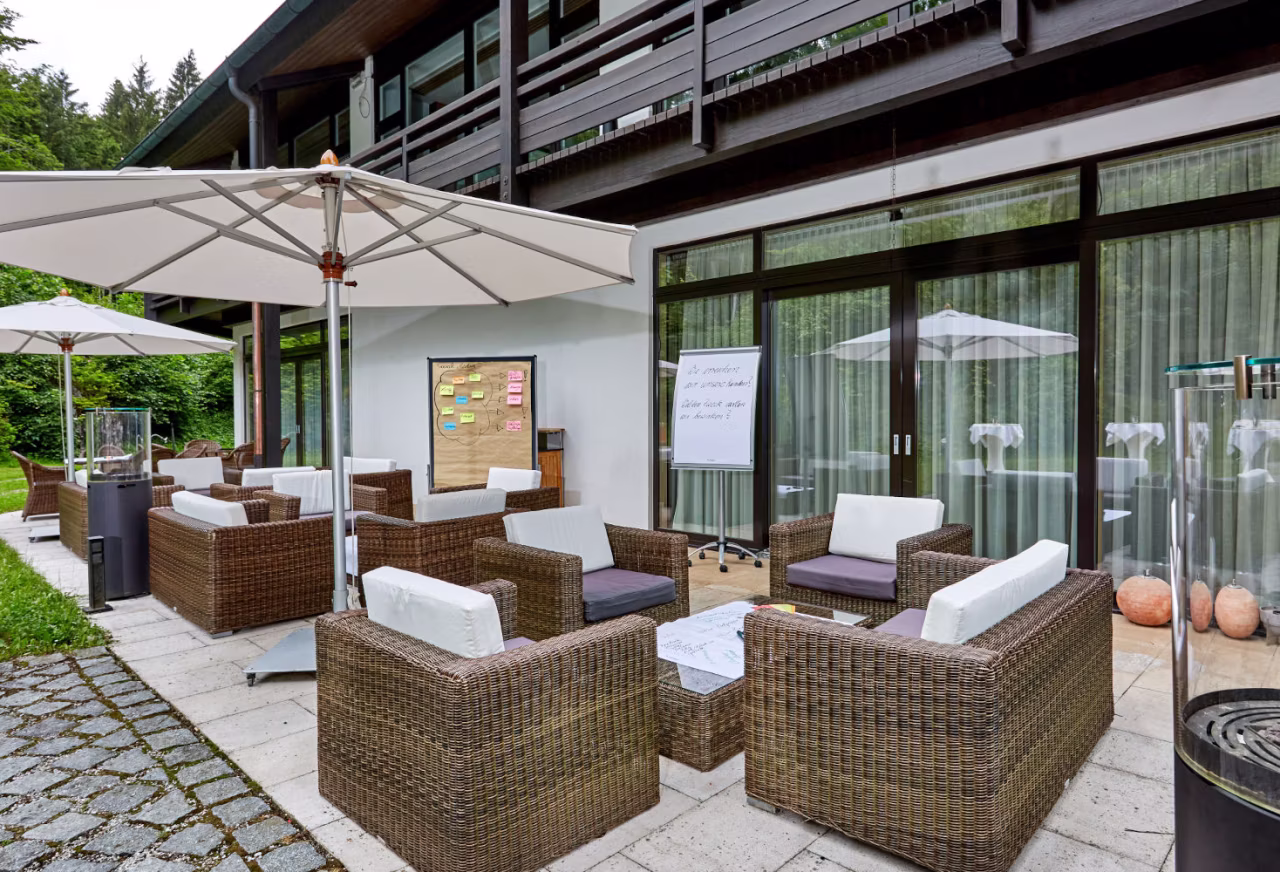 Außenbereich mit Rattansesseln, weißen Sonnenschirmen, Flipchart und großem Glasfensterfront hinter einer modernen Unterkunft.