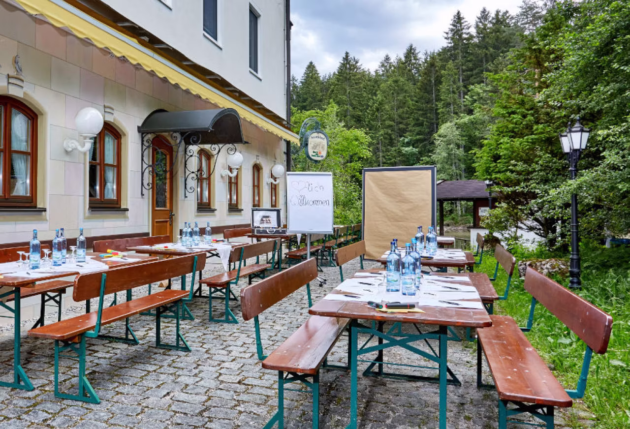 Außenterrasse mit Holzbänken und Tischen, die mit Wasserflaschen und Schreibmaterialien für ein Meeting oder Seminar vorbereitet sind, vor einem Gebäude mit gelbem Sonnenschutz und Wald im Hintergrund.