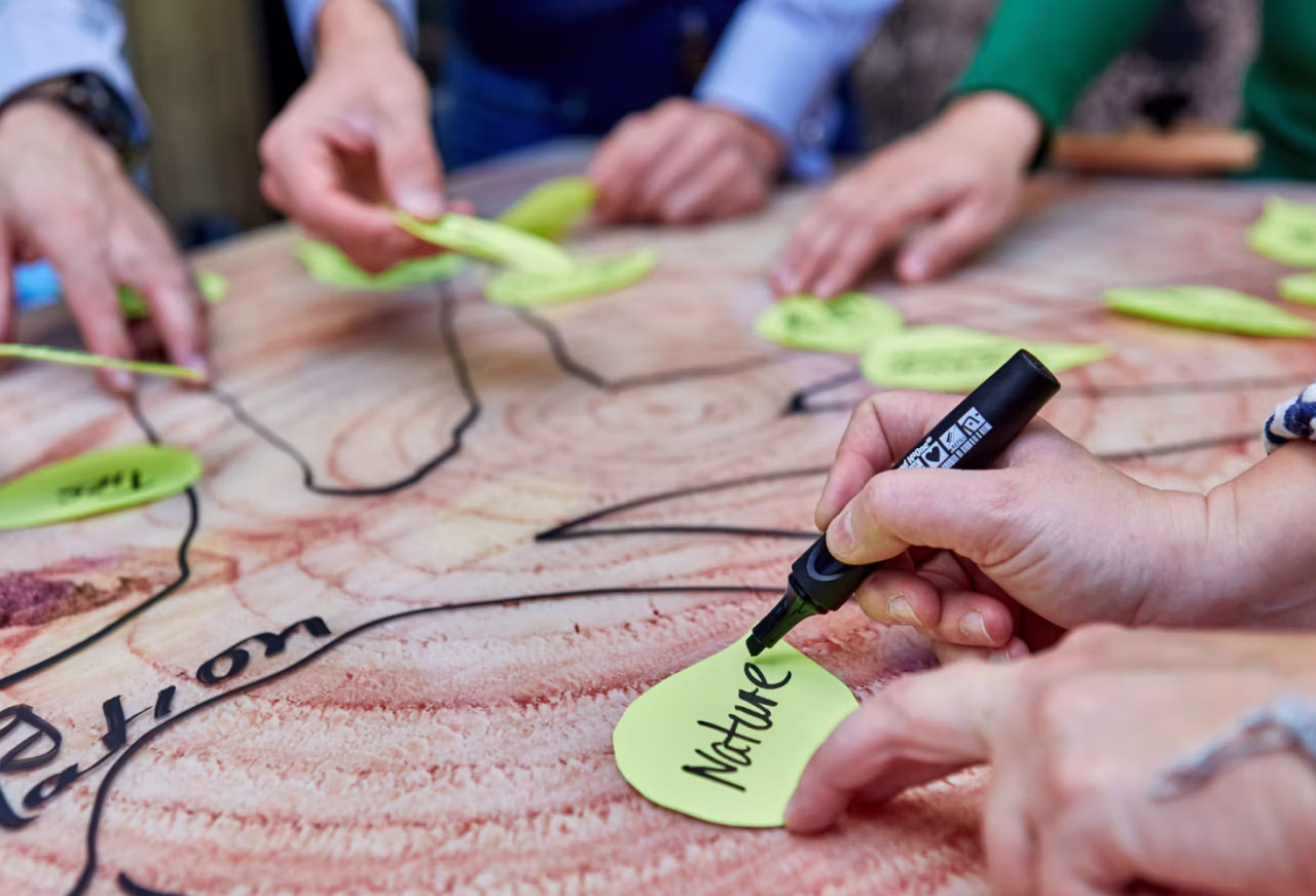 Mehrere Hände legen grüne Notizzettel auf eine hölzerne Oberfläche mit schwarzem Marker, einer schreibt das Wort 'Nature'.
