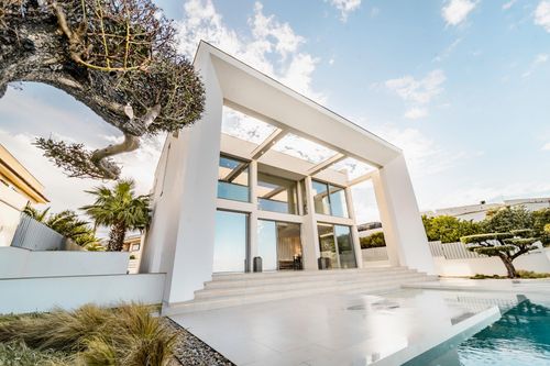 Modern white two-story house with large glass windows, a wide staircase, and a reflective pool in front, surrounded by trees and plants.