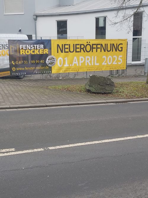 Yellow and black banner on a fence advertising Fenster Rocker with new opening date April 1, 2025, including contact details and website.