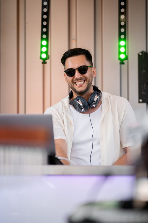 Smiling man wearing sunglasses and headphones around his neck working on a laptop with green lights behind him.