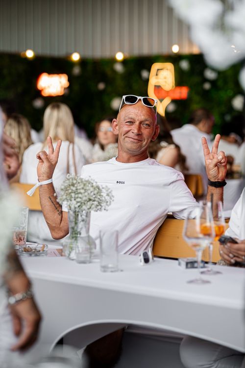 Smiling bald man wearing a white BOSS shirt and sunglasses on his head, making peace signs with both hands at a social event.