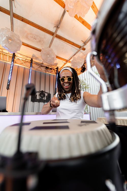 Smiling man with dreadlocks and sunglasses playing conga drums on a lit stage under hanging paper lanterns.