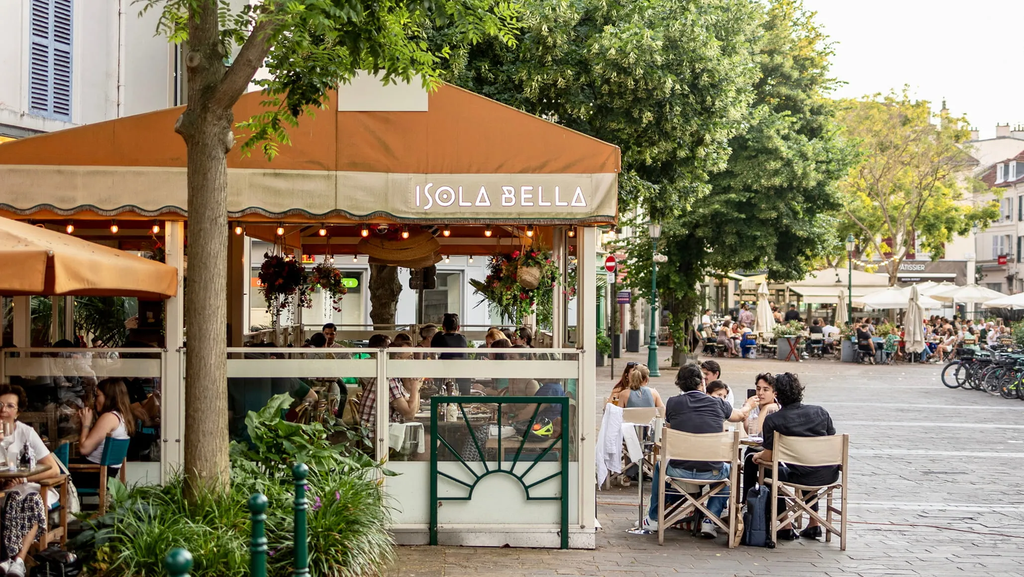 Terrasse de café animée avec des personnes assises sous un auvent orange portant l'inscription Isola Bella, entourée d'arbres et de plantes dans une rue pavée.