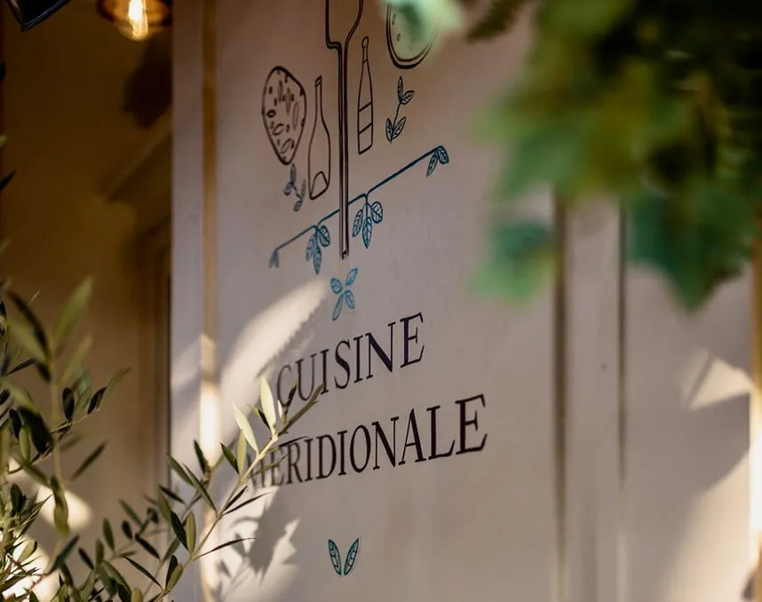 Mur avec inscriptions « CUISINE MÉRIDIONALE » décoré de dessins culinaires et feuillages flous au premier plan.