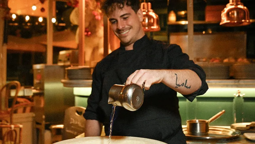 Un chef souriant en tenue noire verse un liquide depuis un petit pot métallique dans une cuisine professionnelle chaleureusement éclairée.