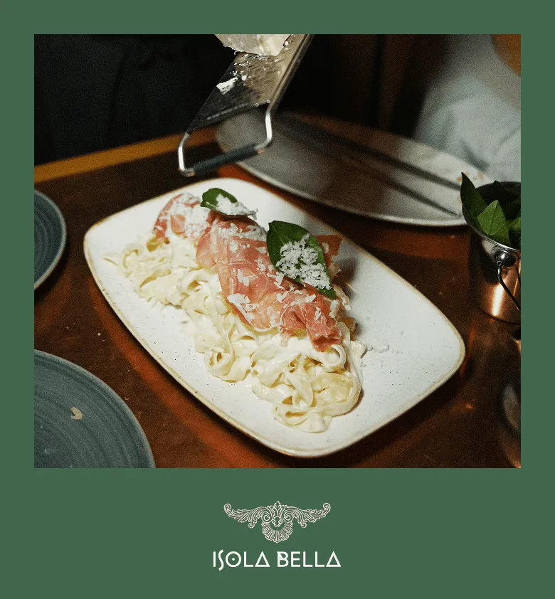 Plat de pâtes crémeuses garnies de tranches de jambon cru et de feuilles de basilic, avec du fromage râpé fraîchement ajouté.