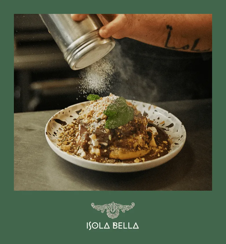 Une main saupoudre du sucre glace sur un dessert garni de sauce au chocolat, de noix concassées et d'une feuille de menthe, sur une assiette blanche à motifs.