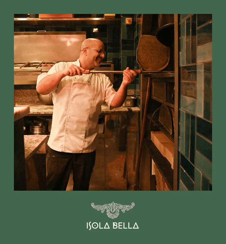 Un chef cuisinier souriant utilisant une pelle en bois pour insérer une pizza dans un four à bois dans une cuisine de pizzeria.