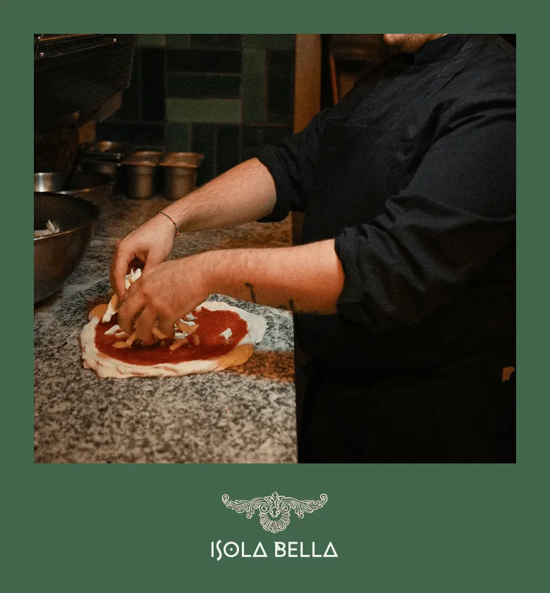 Une personne prépare une pizza en ajoutant du fromage sur une pâte garnie de sauce tomate dans une cuisine.