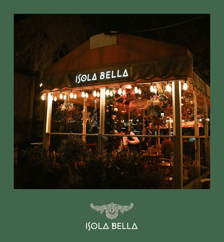 Terrasse extérieure éclairée d'un restaurant nommé Isola Bella avec des guirlandes lumineuses et des décorations florales visibles à travers les vitres.