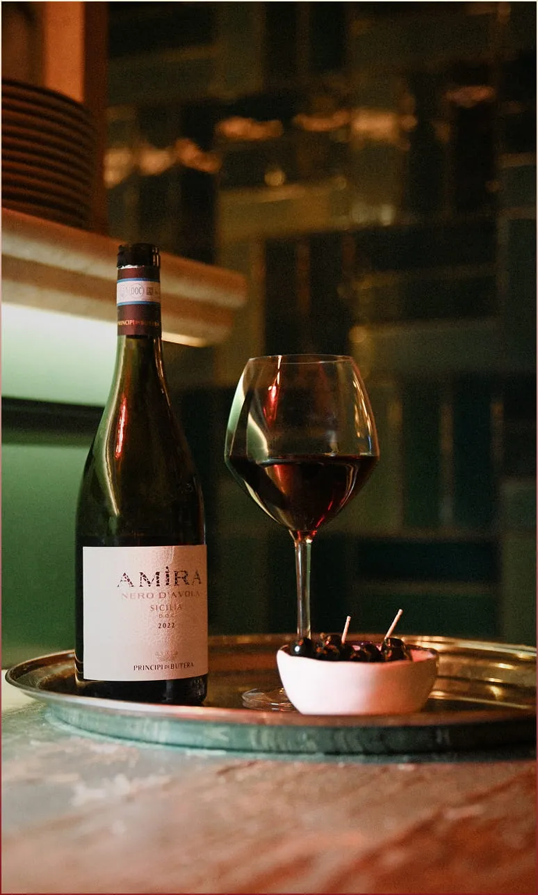 Bouteille de vin rouge Amira Nero d'Avola 2021, verre de vin rempli et petit bol de cerises noires sur un plateau métallique.