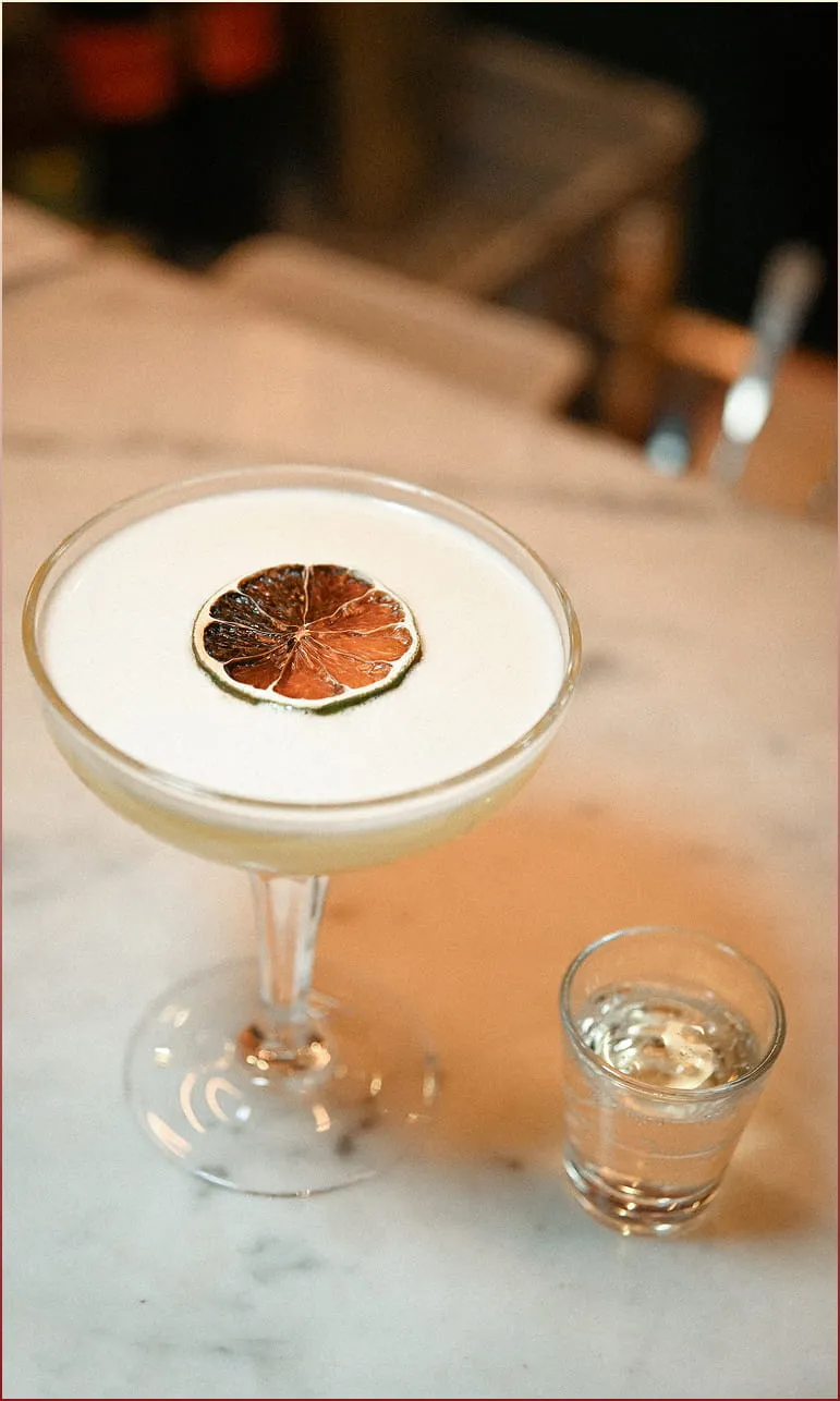 Cocktail mousseux dans un verre à pied avec une tranche de citron séché en garniture, accompagné d'un petit verre d'eau sur une table en marbre.