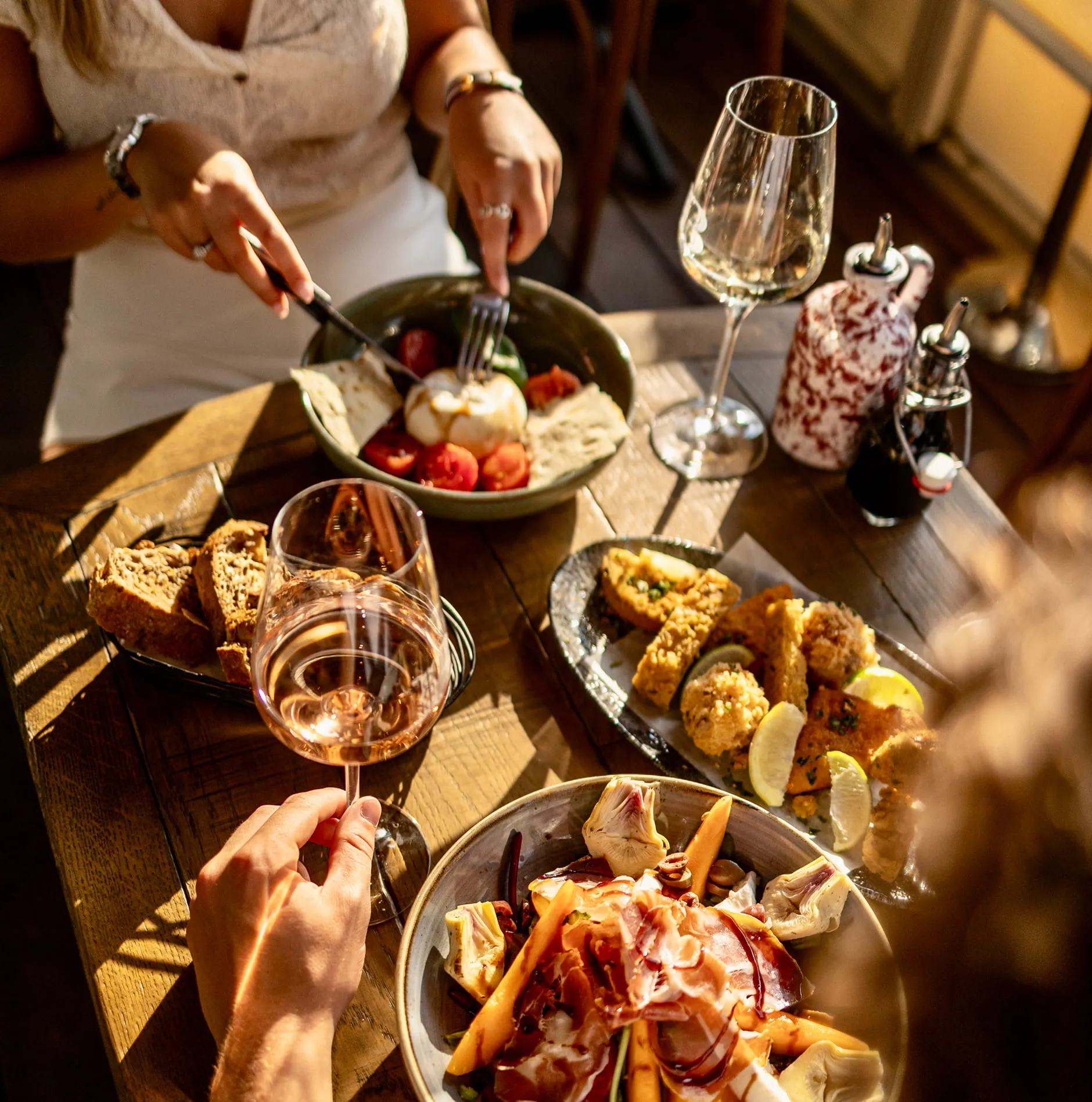 Deux personnes dégustant un repas avec salade, pain, fruits de mer frits, et verre de vin rosé sur une table en bois éclairée par la lumière du soleil.