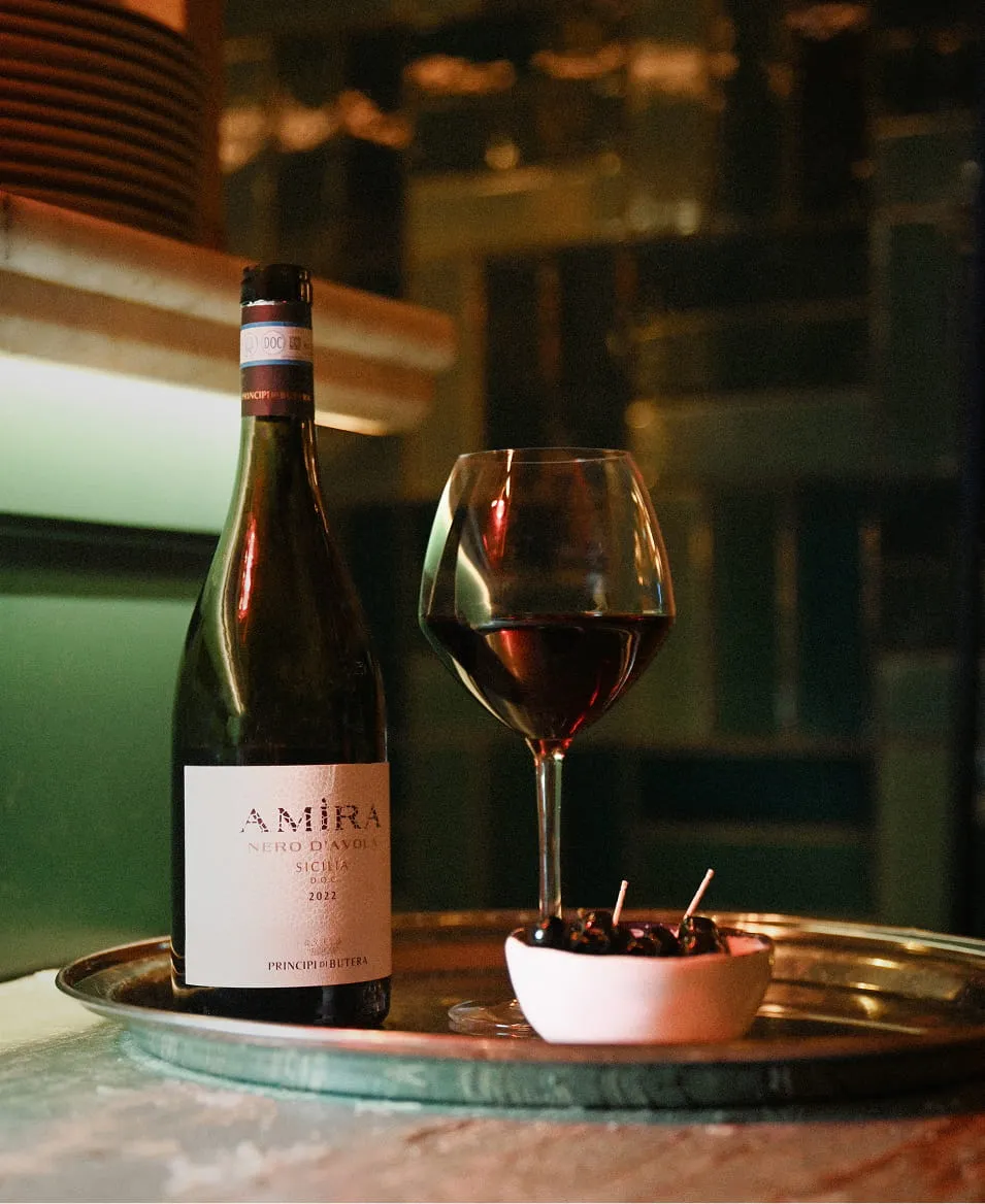 Bouteille de vin rouge Amìra Nero D'Avola 2022 avec un verre de vin et un petit bol de cerises sur un plateau argenté.