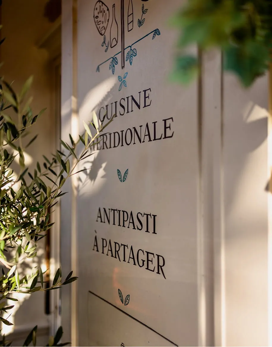 Mur décoré avec texte en français annonçant cuisine méridionale et antipasti à partager, avec des feuilles d'olivier en avant-plan.
