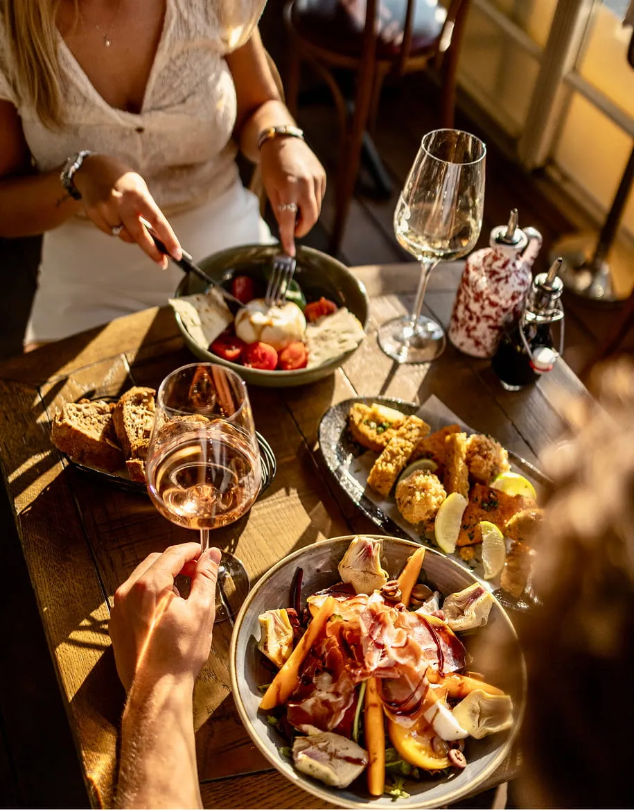 Personnes partageant un repas avec salade, fromage, pain, plats frits et verres de vin sur une table en bois au restaurant.