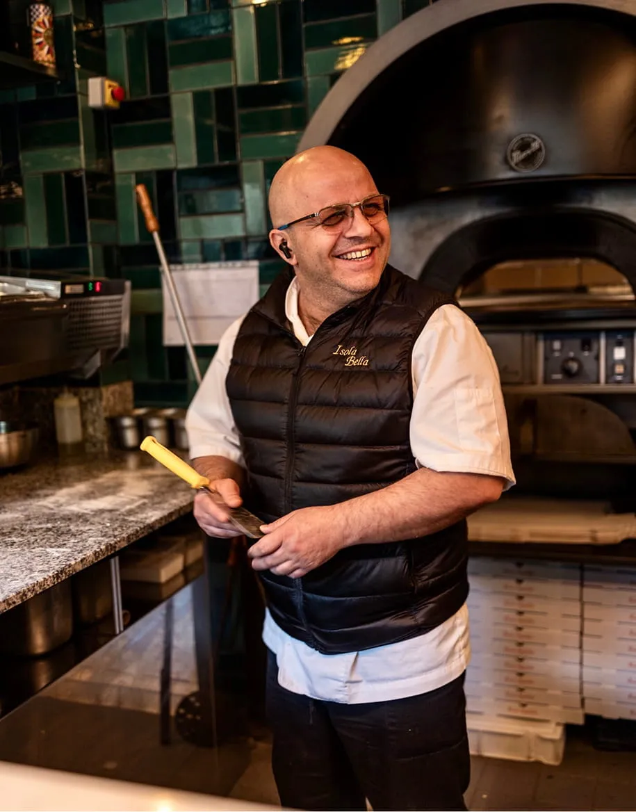Un chef souriant portant un gilet noir 'Isola Bella' dans une cuisine avec un four à pizza en arrière-plan.