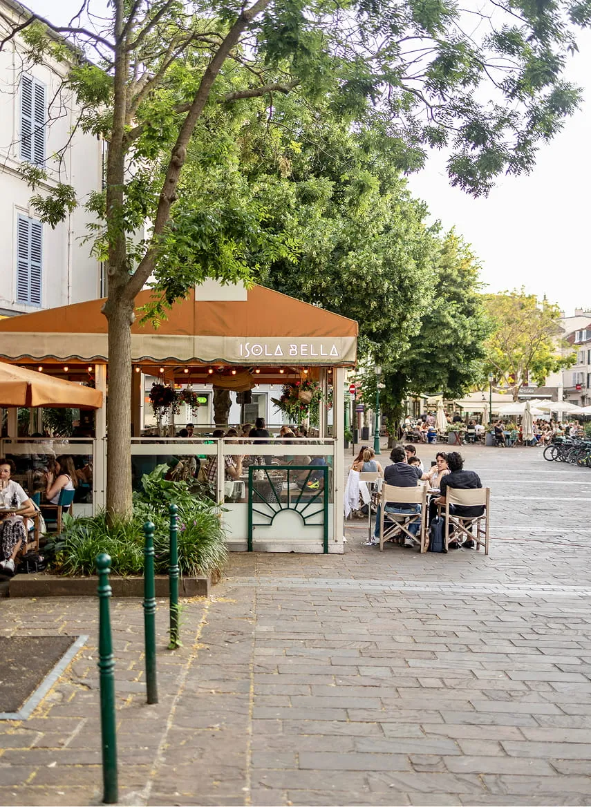 Terrasse animée d'un café nommé Isola Bella avec des clients assis à des tables en plein air sous un auvent orange, entourée d'arbres verts dans une rue pavée.
