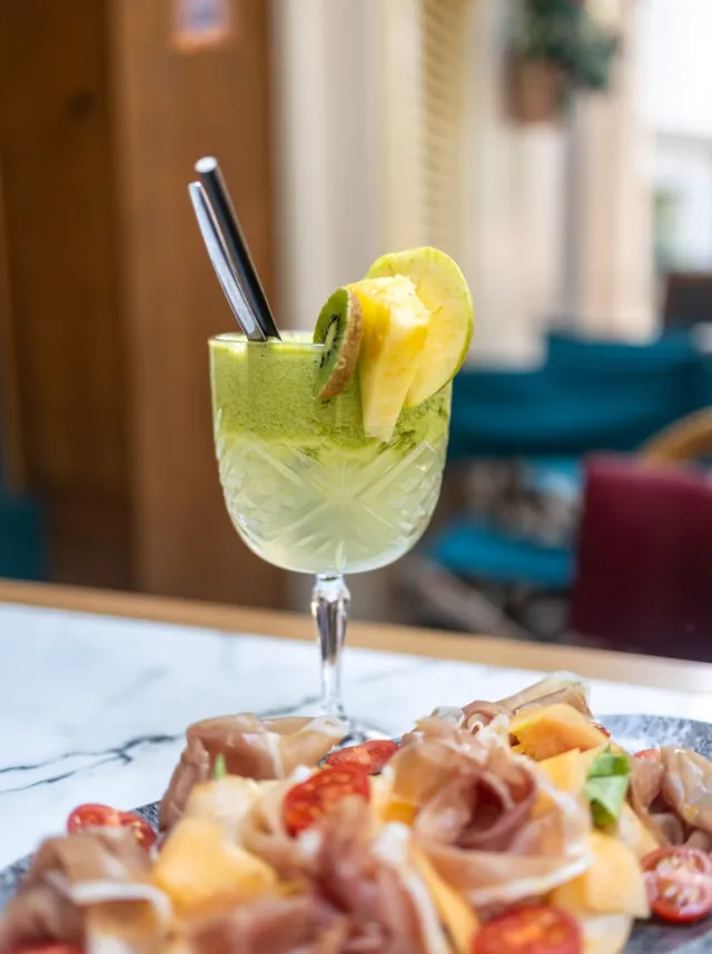 Cocktail vert garnie de tranches de kiwi et d'ananas avec une assiette de charcuterie et tomates cerises en avant-plan.