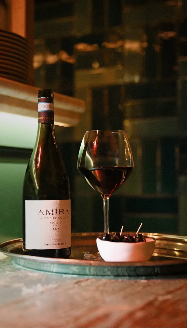 Bouteille de vin rouge Amira Nero d’Avola 2021, verre de vin rouge et petit bol avec olives noires sur un plateau métallique.