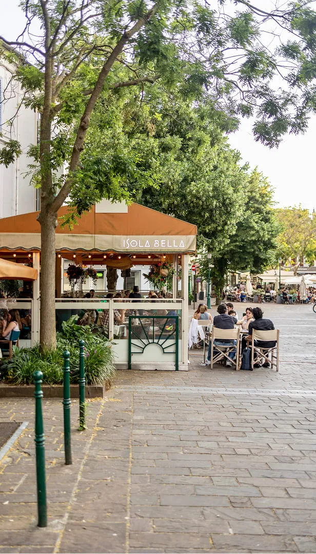 Terrasse de café Isola Bella avec des clients assis à des tables sur une rue pavée, entourée d'arbres verts.