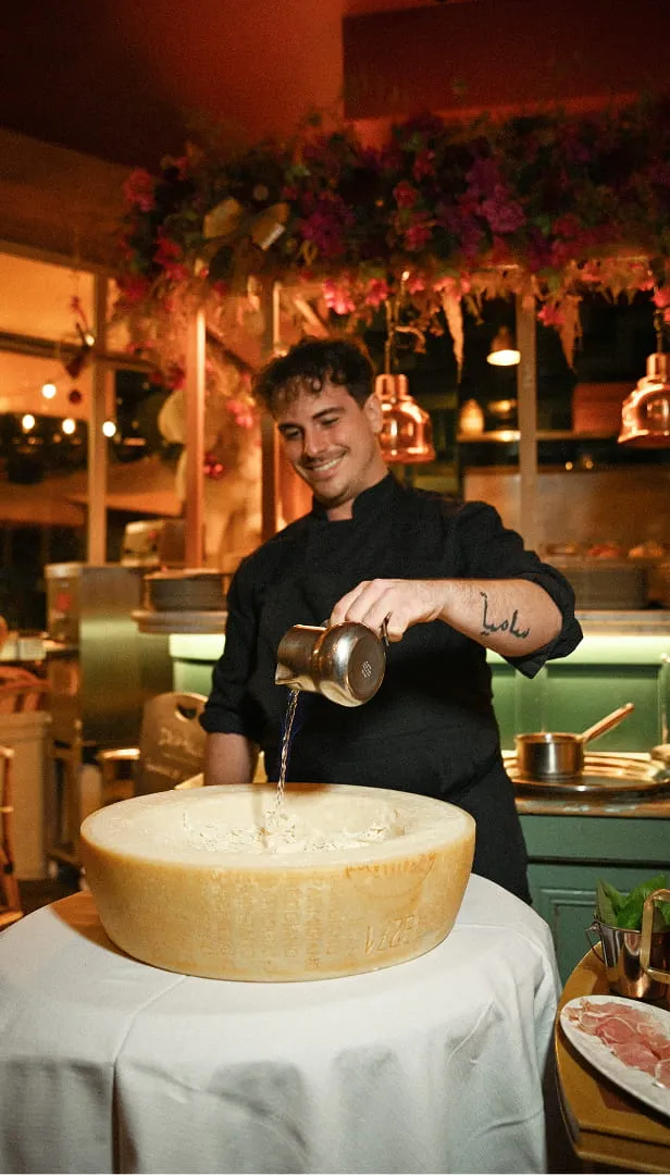 Un chef souriant verse un liquide dans un grand morceau de fromage sur une table ronde dans un restaurant.