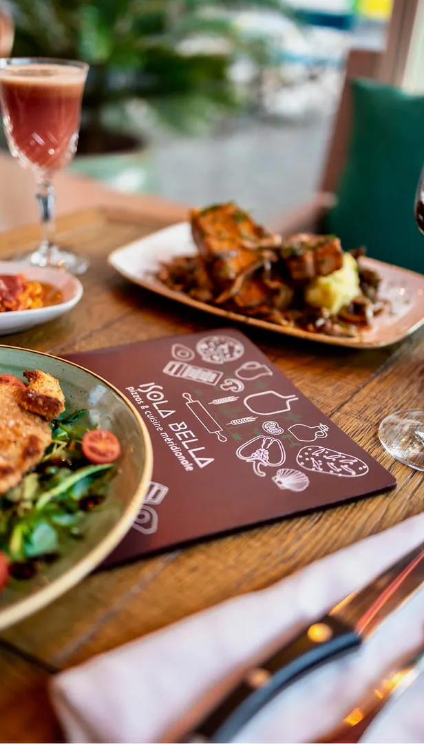 Table en bois avec un menu de restaurant Isola Bella, des assiettes de plats cuisinés et deux verres de vin.