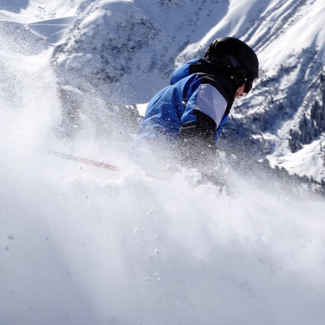 Skier in blue jacket and black helmet skiing downhill with snow spray and snowy mountain backdrop