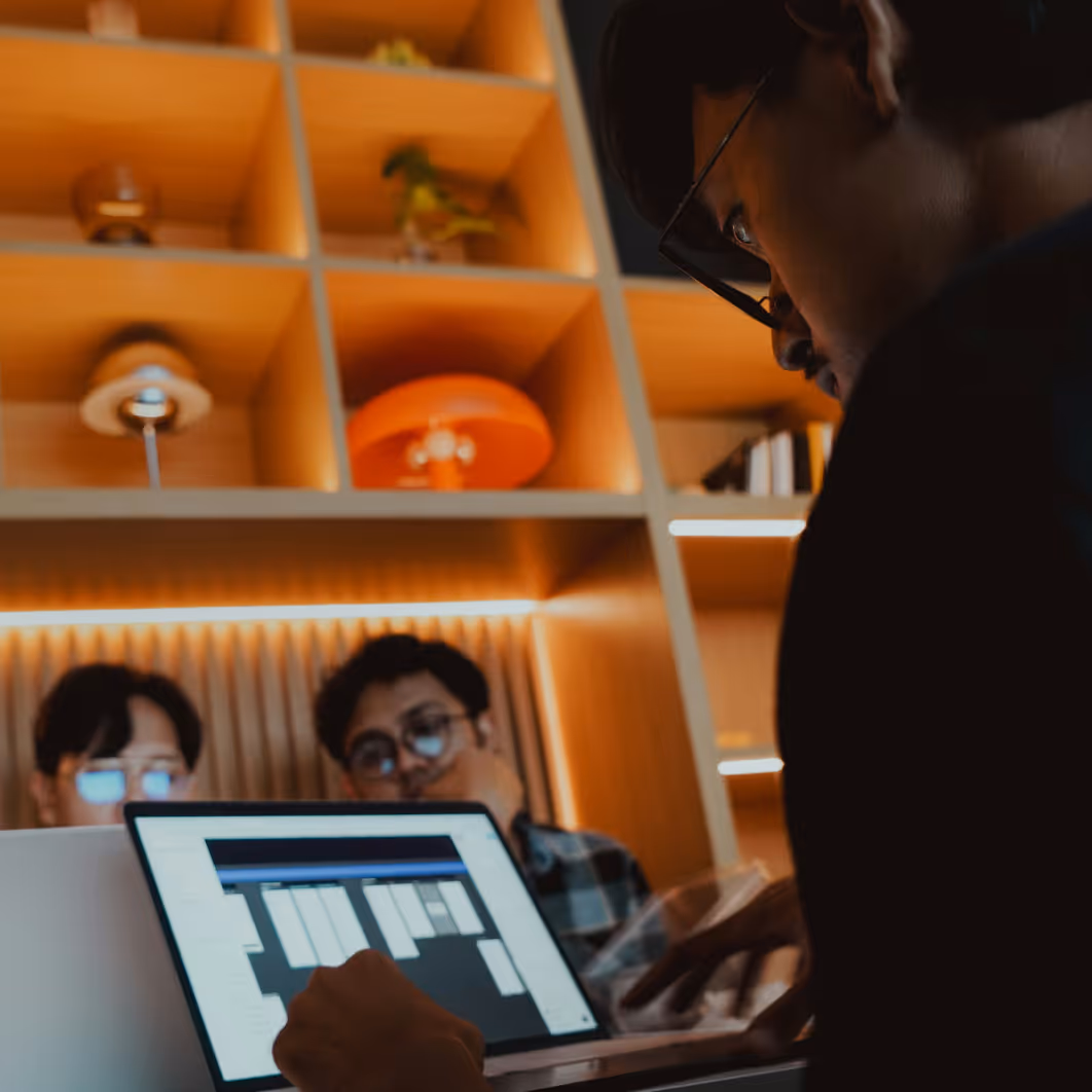 Person focusing on a laptop screen displaying a scheduling or timeline interface with two others in the background
