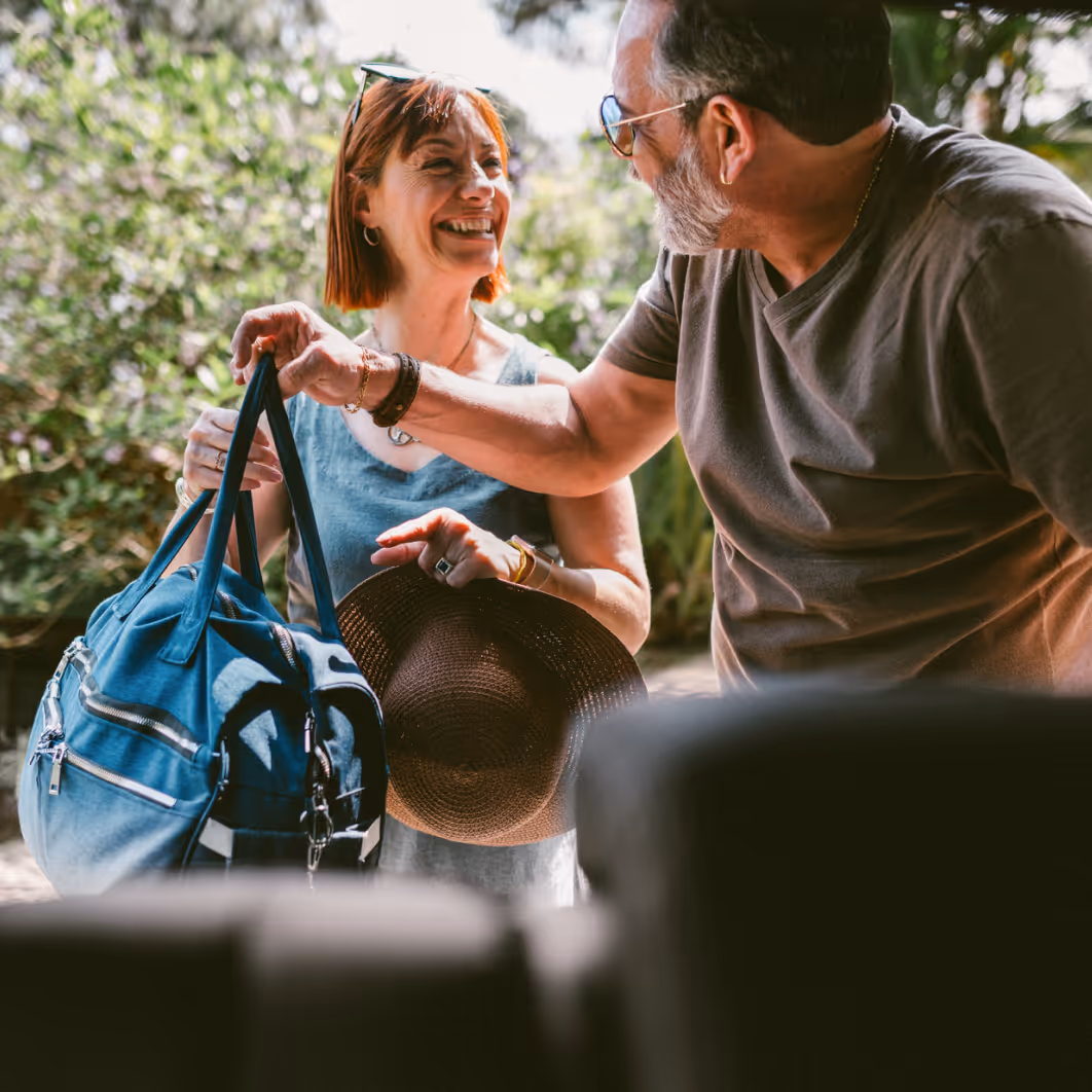 Smiling woman holding a blue travel bag and brown hat while a man reaches to take the bag outdoors