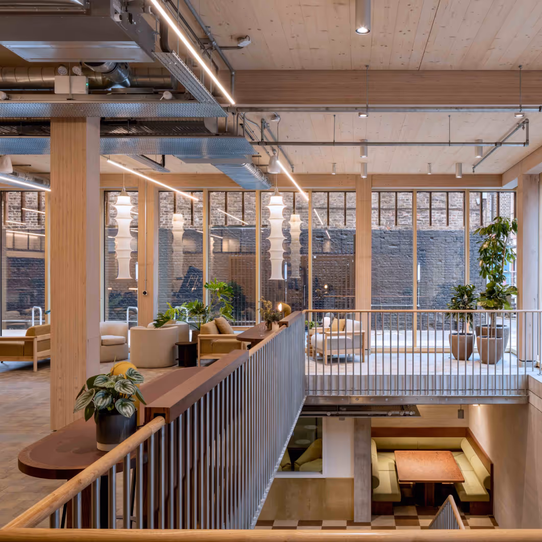 Modern office lounge with natural wood accents, indoor plants, seating areas, and a view of an exposed brick wall