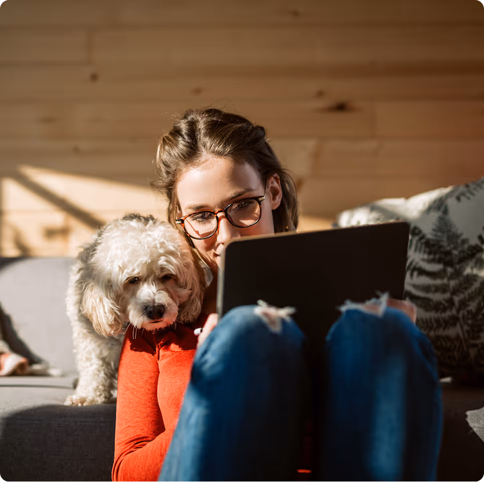 Young woman wearing glasses sitting on a couch using a tablet with a small white dog resting its head on her shoulder
