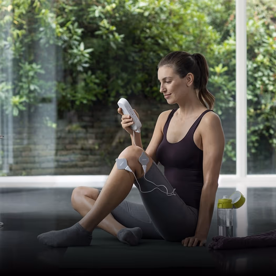Woman sitting on a yoga mat indoors, using an electronic muscle stimulator on her leg, with a water bottle and towel nearby for Omron brand