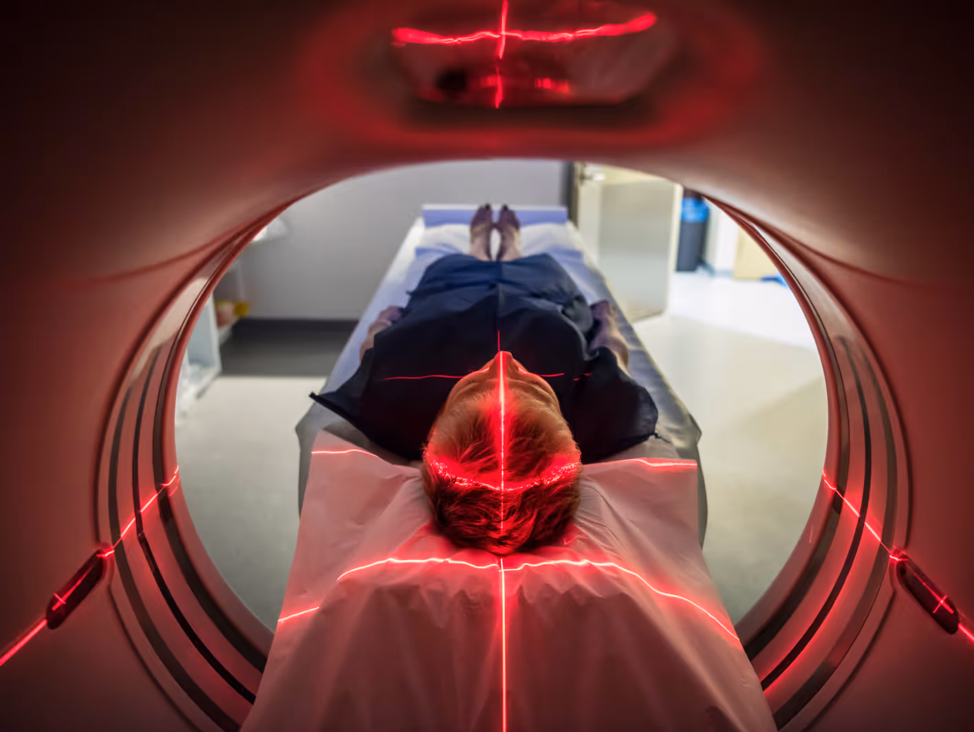 Patient lying on a hospital bed inside an MRI scanner with red laser positioning lines on their head and bed