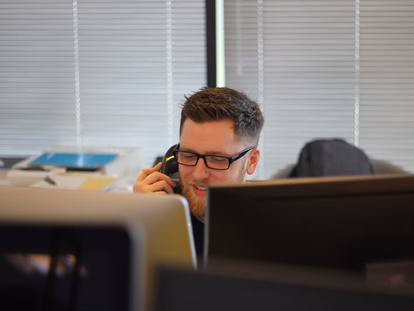 Man with glasses and beard talking on a phone in an office with computer monitors in front of him