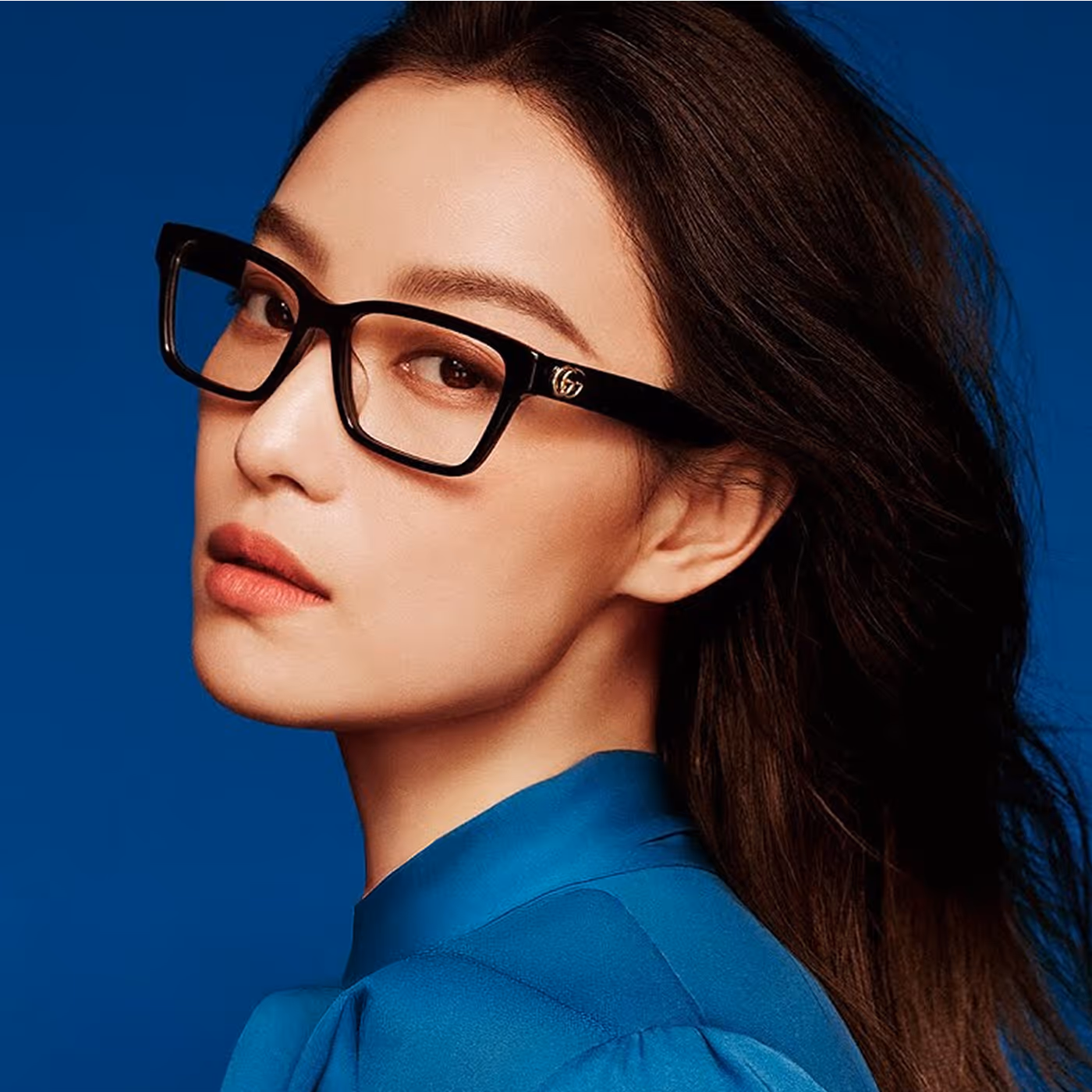 Portrait of a woman with long dark hair wearing black rectangular glasses and a blue blouse against a blue background for Boots Opticians brand