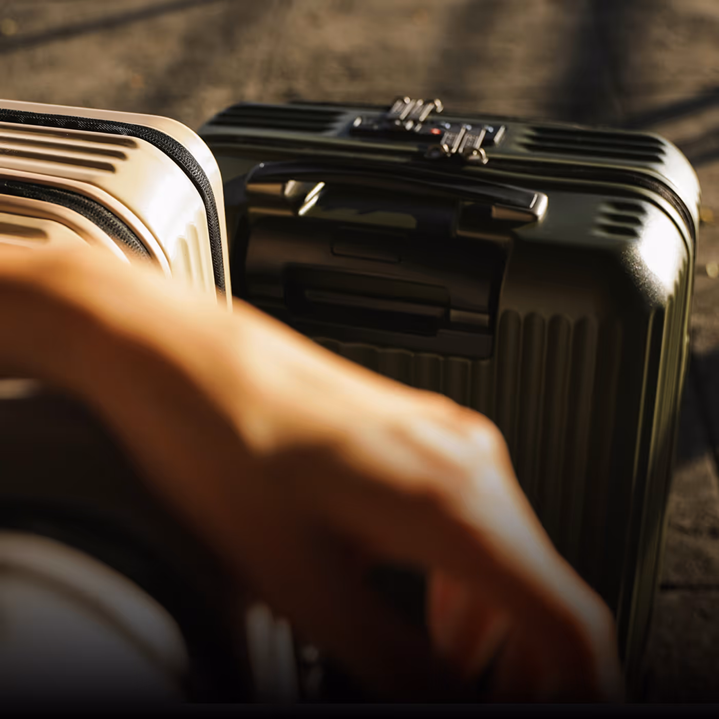 Close-up of two hard-shell suitcases, one beige and one dark green, on a paved surface with a blurred arm in the foreground for Engine brand