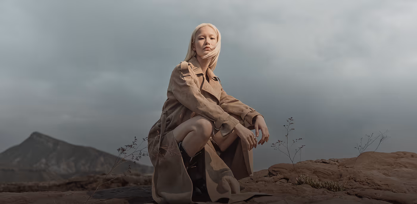 Woman in beige trench coat and black boots crouching on rocky terrain with mountain and cloudy sky in the background for AllSaints brand